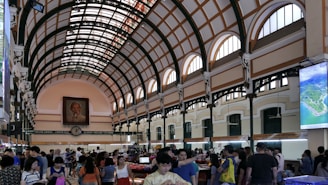 A bustling interior of a large postal building with a high arched ceiling featuring a grid pattern of orange and cream. Numerous people are milling about, browsing items at stalls. A large portrait is prominently hung on the far end wall above an antique-style clock. Daylight streams through the arched windows, illuminating the space.