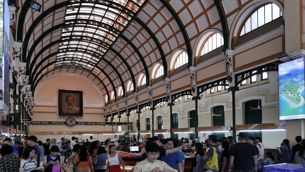 A bustling interior of a large postal building with a high arched ceiling featuring a grid pattern of orange and cream. Numerous people are milling about, browsing items at stalls. A large portrait is prominently hung on the far end wall above an antique-style clock. Daylight streams through the arched windows, illuminating the space.