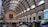 A bustling interior of a large postal building with a high arched ceiling featuring a grid pattern of orange and cream. Numerous people are milling about, browsing items at stalls. A large portrait is prominently hung on the far end wall above an antique-style clock. Daylight streams through the arched windows, illuminating the space.