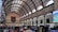 A bustling interior of a large postal building with a high arched ceiling featuring a grid pattern of orange and cream. Numerous people are milling about, browsing items at stalls. A large portrait is prominently hung on the far end wall above an antique-style clock. Daylight streams through the arched windows, illuminating the space.