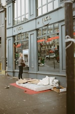 A person is using an ATM outside a building with the logo LCL on it. On the sidewalk, there is a mattress with some cardboard and bedding, suggesting a makeshift sleeping area. Reflections can be seen in the large windows of the building.