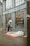 A person is using an ATM outside a building with the logo LCL on it. On the sidewalk, there is a mattress with some cardboard and bedding, suggesting a makeshift sleeping area. Reflections can be seen in the large windows of the building.