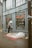A person is using an ATM outside a building with the logo LCL on it. On the sidewalk, there is a mattress with some cardboard and bedding, suggesting a makeshift sleeping area. Reflections can be seen in the large windows of the building.