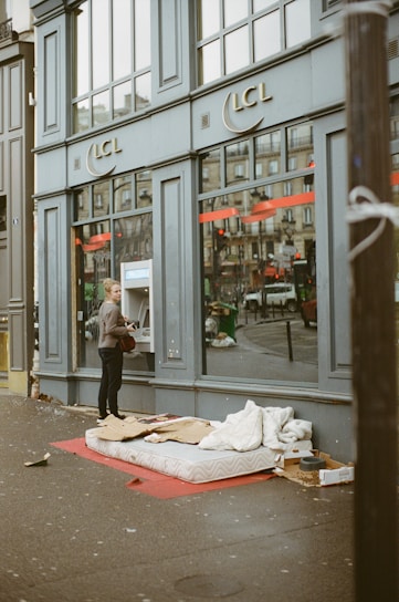 A person is using an ATM outside a building with the logo LCL on it. On the sidewalk, there is a mattress with some cardboard and bedding, suggesting a makeshift sleeping area. Reflections can be seen in the large windows of the building.
