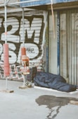 A sleeping bag lies on the sidewalk next to a closed shop with metal shutters covered in graffiti. There is scaffolding nearby, along with a shopping cart filled with personal items and a poster on a pole. A wet patch is visible on the pavement.