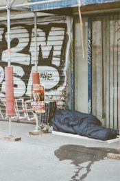 A sleeping bag lies on the sidewalk next to a closed shop with metal shutters covered in graffiti. There is scaffolding nearby, along with a shopping cart filled with personal items and a poster on a pole. A wet patch is visible on the pavement.