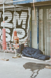 A sleeping bag lies on the sidewalk next to a closed shop with metal shutters covered in graffiti. There is scaffolding nearby, along with a shopping cart filled with personal items and a poster on a pole. A wet patch is visible on the pavement.