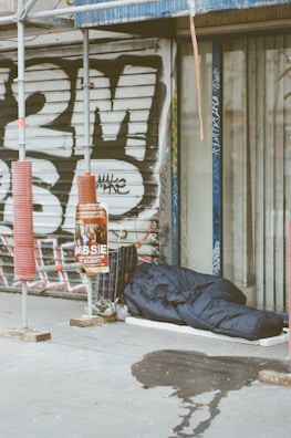 A sleeping bag lies on the sidewalk next to a closed shop with metal shutters covered in graffiti. There is scaffolding nearby, along with a shopping cart filled with personal items and a poster on a pole. A wet patch is visible on the pavement.