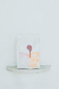 A minimalist packaging bag with a graphic design in light pink and orange hues. The print includes abstract shapes and a tree, with a circular red seal at the top center.