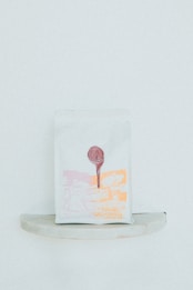 A minimalist packaging bag with a graphic design in light pink and orange hues. The print includes abstract shapes and a tree, with a circular red seal at the top center.