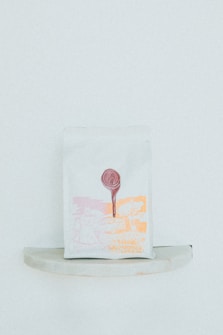 A minimalist packaging bag with a graphic design in light pink and orange hues. The print includes abstract shapes and a tree, with a circular red seal at the top center.