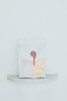 A minimalist packaging bag with a graphic design in light pink and orange hues. The print includes abstract shapes and a tree, with a circular red seal at the top center.