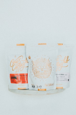 Three white coffee bags with detailed golden and red labels displaying artistic designs and text, including a skull motif on the central bag.