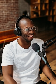 A person wearing glasses and headphones is smiling while speaking into a microphone. The setting appears to be a recording studio with a brick wall and wooden shelves in the background. They are seated comfortably, engaging in what might be a podcast or audio recording session.