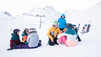 Several people wearing colorful winter clothing are gathered on a snowy slope, surrounded by snowboards and ski gear. A person in a yellow jacket kneels while speaking into a microphone, seemingly conducting an interview. The backdrop features majestic snow-covered mountains and a ski lift in the distance, creating a serene yet lively winter scene.