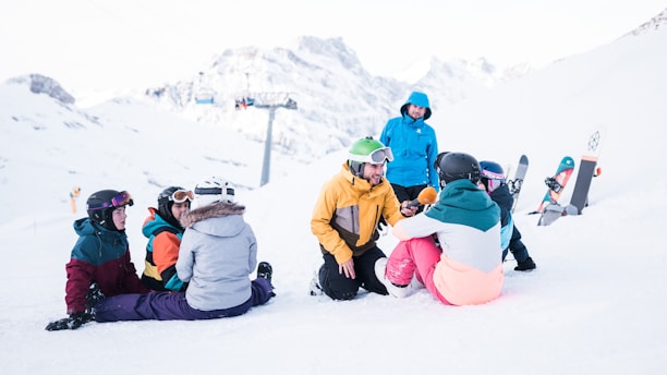 Several people wearing colorful winter clothing are gathered on a snowy slope, surrounded by snowboards and ski gear. A person in a yellow jacket kneels while speaking into a microphone, seemingly conducting an interview. The backdrop features majestic snow-covered mountains and a ski lift in the distance, creating a serene yet lively winter scene.