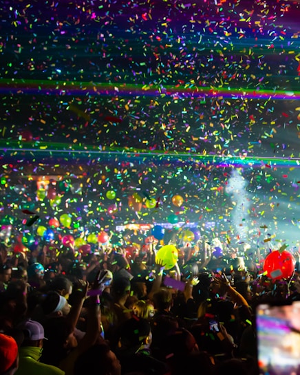 A vibrant crowd enjoying a lively concert or festival setting, surrounded by colorful confetti and party lights. Many people hold up their hands, capturing the moment with phones while bright balloons float above them.
