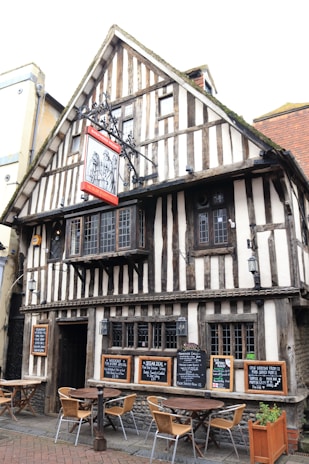 A half-timbered building with a steep gabled roof and Tudor-style architecture displays several blackboards advertizing food menus. The ground level has wooden tables and chairs arranged outside. The upper part of the building features ornate metalwork signage and a set of small, multi-pane windows.