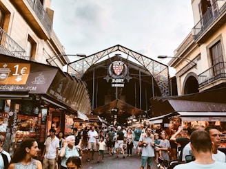 A bustling market scene with numerous people walking and shopping under a large, ornate entrance structure. The market is lined with stalls displaying various goods. The entrance sign reads 'Mercat St. Josep La Boqueria'. Surrounding the market are buildings with balconies.