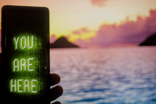 A smartphone displaying a neon green message 'YOU ARE HERE' is held up in the foreground against a serene seascape backdrop. The sky is painted in vibrant hues of pink, orange, and purple as the sun sets behind distant hills, with calm waters reflecting the colorful sky.
