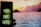A smartphone displaying a neon green message 'YOU ARE HERE' is held up in the foreground against a serene seascape backdrop. The sky is painted in vibrant hues of pink, orange, and purple as the sun sets behind distant hills, with calm waters reflecting the colorful sky.