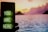 A smartphone displaying a neon green message 'YOU ARE HERE' is held up in the foreground against a serene seascape backdrop. The sky is painted in vibrant hues of pink, orange, and purple as the sun sets behind distant hills, with calm waters reflecting the colorful sky.