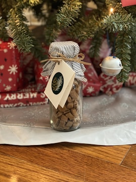 A festive scene with a glass jar filled with brown sugar cubes, adorned with a striped fabric cover and a decorative tag featuring a pine motif. The jar is placed beneath a Christmas tree, surrounded by wrapped gifts in red paper with white star patterns and a silver bell ornament hanging from the tree.