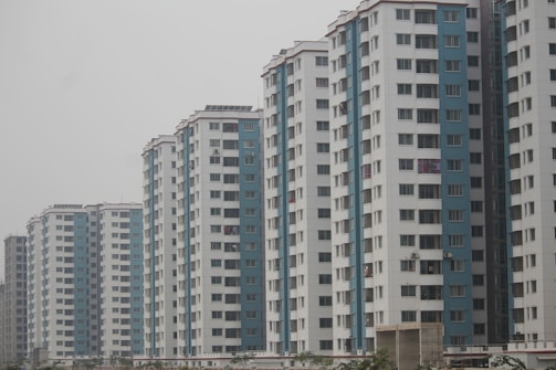 Several high-rise apartment buildings are aligned in a row. Each building has a consistent design with alternating white and blue vertical sections. Numerous windows are visible on each floor, and some balconies display laundry or other items. The sky is overcast, giving a muted appearance to the scene.