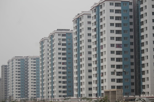 Several high-rise apartment buildings are aligned in a row. Each building has a consistent design with alternating white and blue vertical sections. Numerous windows are visible on each floor, and some balconies display laundry or other items. The sky is overcast, giving a muted appearance to the scene.