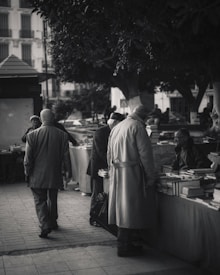 A black and white image of an outdoor market scene where several people are gathered around tables filled with books. The people are wearing coats, suggesting cool weather. Large trees provide shade and create a cozy atmosphere. Buildings are visible in the background.
