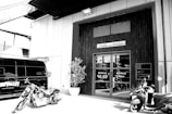 A monochrome image of an industrial-style building with a sign that says Pine Valley above the entrance. There's a motorcycle and a scooter parked in front of the entrance, and a dark van with branding on the side. A potted plant is situated near the doorway.