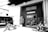 A monochrome image of an industrial-style building with a sign that says Pine Valley above the entrance. There's a motorcycle and a scooter parked in front of the entrance, and a dark van with branding on the side. A potted plant is situated near the doorway.