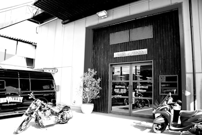 A monochrome image of an industrial-style building with a sign that says Pine Valley above the entrance. There's a motorcycle and a scooter parked in front of the entrance, and a dark van with branding on the side. A potted plant is situated near the doorway.