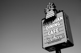 A rectangular signboard with an image of a pineapple at the top promotes a Hawaiian dining cafe. The text on the sign reads 'Hawaiian Dining Cafe La Ohana' in bold letters. The sign is set against a clear sky.