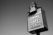 A rectangular signboard with an image of a pineapple at the top promotes a Hawaiian dining cafe. The text on the sign reads 'Hawaiian Dining Cafe La Ohana' in bold letters. The sign is set against a clear sky.