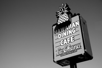 A rectangular signboard with an image of a pineapple at the top promotes a Hawaiian dining cafe. The text on the sign reads 'Hawaiian Dining Cafe La Ohana' in bold letters. The sign is set against a clear sky.