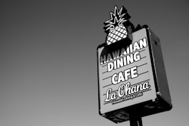 A rectangular signboard with an image of a pineapple at the top promotes a Hawaiian dining cafe. The text on the sign reads 'Hawaiian Dining Cafe La Ohana' in bold letters. The sign is set against a clear sky.