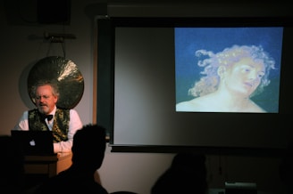 A man stands at a podium with a laptop in front of him, delivering a lecture or presentation. Behind him, there is a large gong on the wall. To his right, a projector displays an artwork featuring a head and shoulders portrait of a person with curly hair, set against a dark background. The room is dimly lit, giving the scene a focused, academic atmosphere.