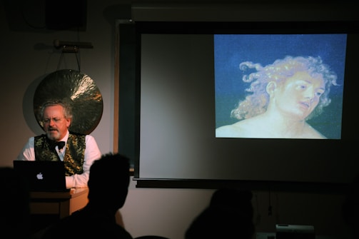 A man stands at a podium with a laptop in front of him, delivering a lecture or presentation. Behind him, there is a large gong on the wall. To his right, a projector displays an artwork featuring a head and shoulders portrait of a person with curly hair, set against a dark background. The room is dimly lit, giving the scene a focused, academic atmosphere.