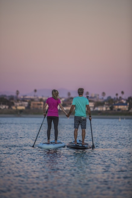 A couple paddleboarding side by side with the sun setting behind the calm ocean horizon.