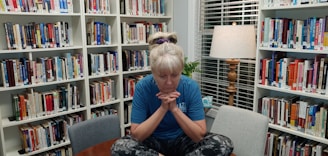 A person with blond hair is sitting cross-legged on a small table in a library, surrounded by bookshelves filled with various books. The individual is wearing a blue shirt and patterned leggings, with a pensive expression, supporting their chin with their hands. A lamp with a wooden base and a fabric shade is lit in the background, casting a warm light. There is a small plant and a decorative mug near the window with blinds.