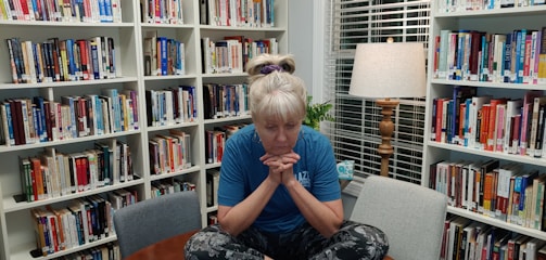 A person with blond hair is sitting cross-legged on a small table in a library, surrounded by bookshelves filled with various books. The individual is wearing a blue shirt and patterned leggings, with a pensive expression, supporting their chin with their hands. A lamp with a wooden base and a fabric shade is lit in the background, casting a warm light. There is a small plant and a decorative mug near the window with blinds.