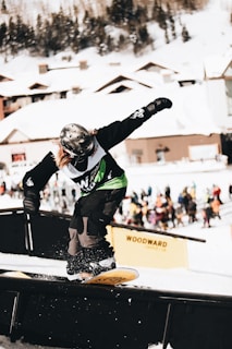 A snowboarder wearing a helmet and green bib performs a trick on a rail in a snowy resort setting. The background features snow-covered wooden cabins and blurred spectators.