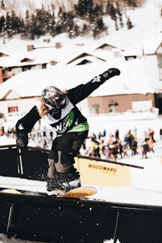 A snowboarder wearing a helmet and green bib performs a trick on a rail in a snowy resort setting. The background features snow-covered wooden cabins and blurred spectators.