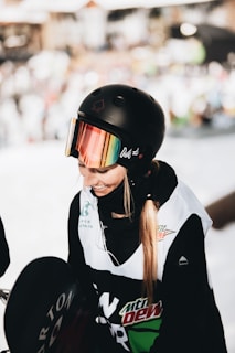 A person is wearing a black helmet with reflective goggles and a black jacket adorned with various logos. The background is blurred, suggesting an outdoor winter environment.