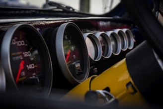 A close-up view of multiple car dashboard gauges, including speedometers and tachometers, with reflections visible on their glass surfaces. The gauges have white borders and are set against a glossy black dashboard. Some of the meters display numbers, needles, and indicators in red and white.