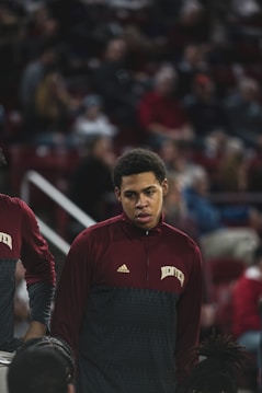 A person wearing a maroon and gray athletic jacket with the word 'DENVER' across the chest stands in an indoor sports arena. The background is filled with blurred spectators seated in rows, creating a sense of anticipation and focus on the individual.