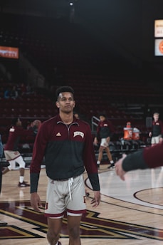 A basketball player wearing a maroon and white uniform stands on a basketball court, with other players in the background. The arena appears dimly lit, with some spectators visible in the stands. The player is holding a basketball and seems focused.