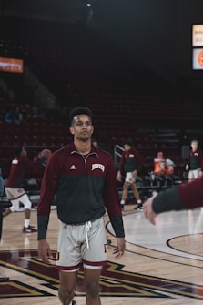 A basketball player wearing a maroon and white uniform stands on a basketball court, with other players in the background. The arena appears dimly lit, with some spectators visible in the stands. The player is holding a basketball and seems focused.