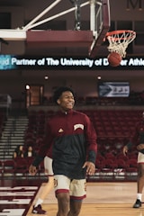 A basketball player in a maroon and white uniform smiles while walking on a basketball court. The ball is mid-air near the hoop, indicating a successful shot. In the background, there are empty red stands and a digital banner displaying text related to the University of Denver Athletics.